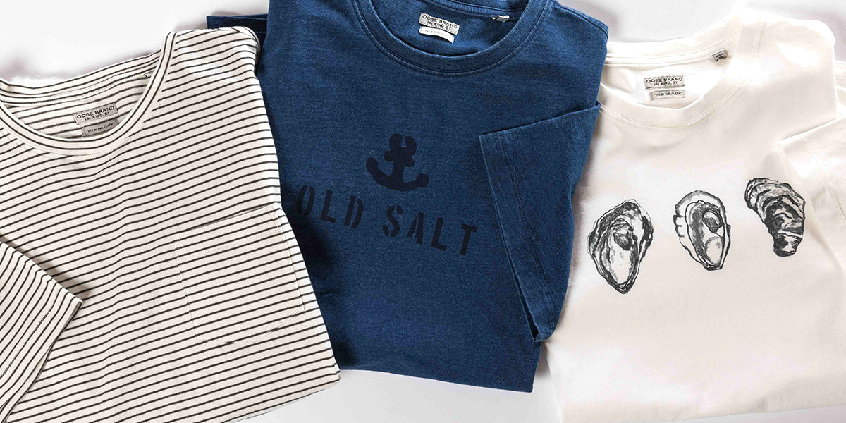 Three OOBE Brand t-shirts folded and overlapping on a table. Far left t-shirt is white and navy stripe, middle t-shirt is navy with an anchor graphic that says Old Salt. The third t-shirt is white with oyster graphic across the top center in black.